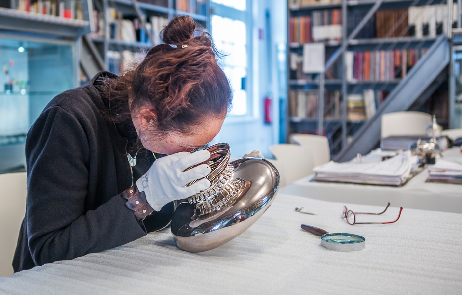 Vrouw met handschoenen aan bekijkt zilveren schaal, met loep en bril op tafel