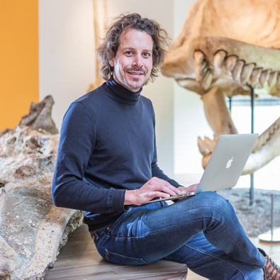 Man op laptop in museum, dinosaurus schedel op de achtergrond