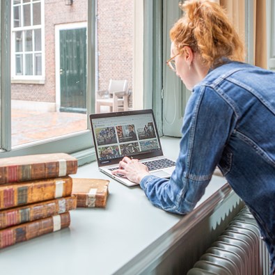 Thema digitalisering: afbeelding van een vrouw met laptop en een stapel oude boeken in een vensterbank