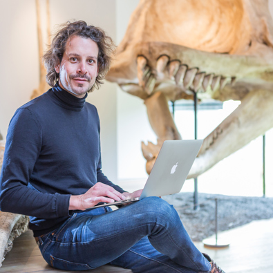 Man zit met laptop op schoot met op achtergrond skelet van dinosaurus