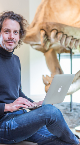 Man zit met laptop op schoot met op achtergrond skelet van dinosaurus