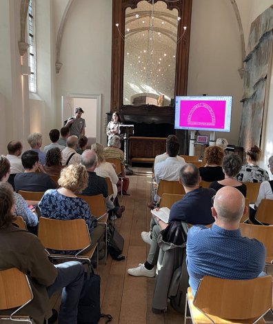 Foto achterin de Van Assendelftkapel gericht op het podium en publiek, de zaal waar de bijeenkomst gehouden werd.