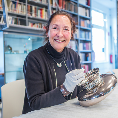 Vrouw zit met handschoenen aan tafel bij zilveren object, met loep en bril op tafel