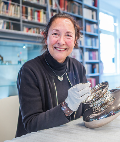 Vrouw zit met handschoenen aan tafel bij zilveren object, met loep en bril op tafel