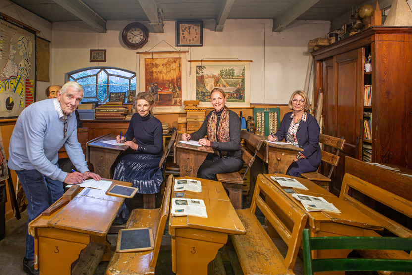 Man en drie vrouwen in ouderwets klaslokaal, man ondertekend document.